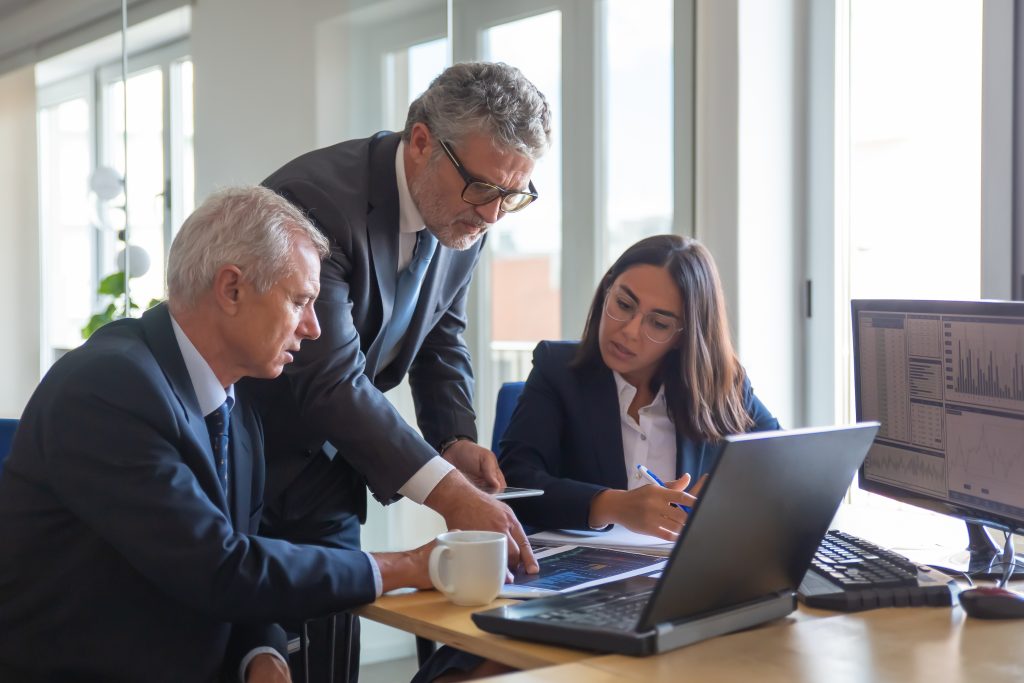 three business professionals gather to look at information on a laptop in an office conference room.