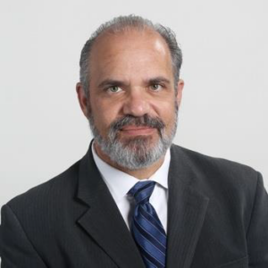 Richard Baratta PhD wears a grey suit and blue tie. He has neatly trimmed facial hair and smiles at the camera for his head-shot.