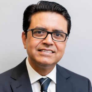 A photograph of Mohammed Mushtaq. He wears a dark suit, tie, and thick-rimmed glasses. He smiles at the camera.
