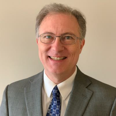 A photograph of Timothy Rhoades PhD in a light grey suit and blue tie. He wears thin rimmed glasses and smiles at the camera.