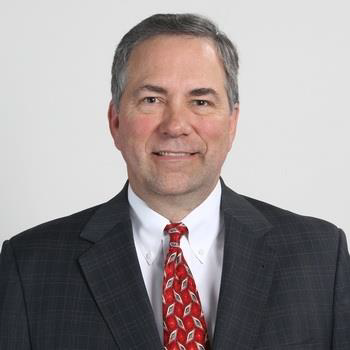 A photograph of Dirk Smith PhD wearing a grey suit with a red tie.
