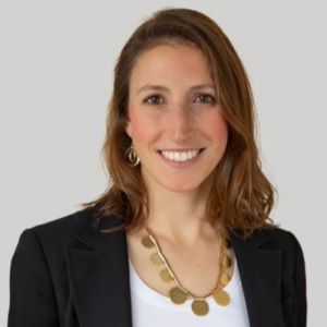 A headshot of Jessica Zendler PhD wearing a black suit jacket over a white t-shirt with a gold necklace and earrrings.